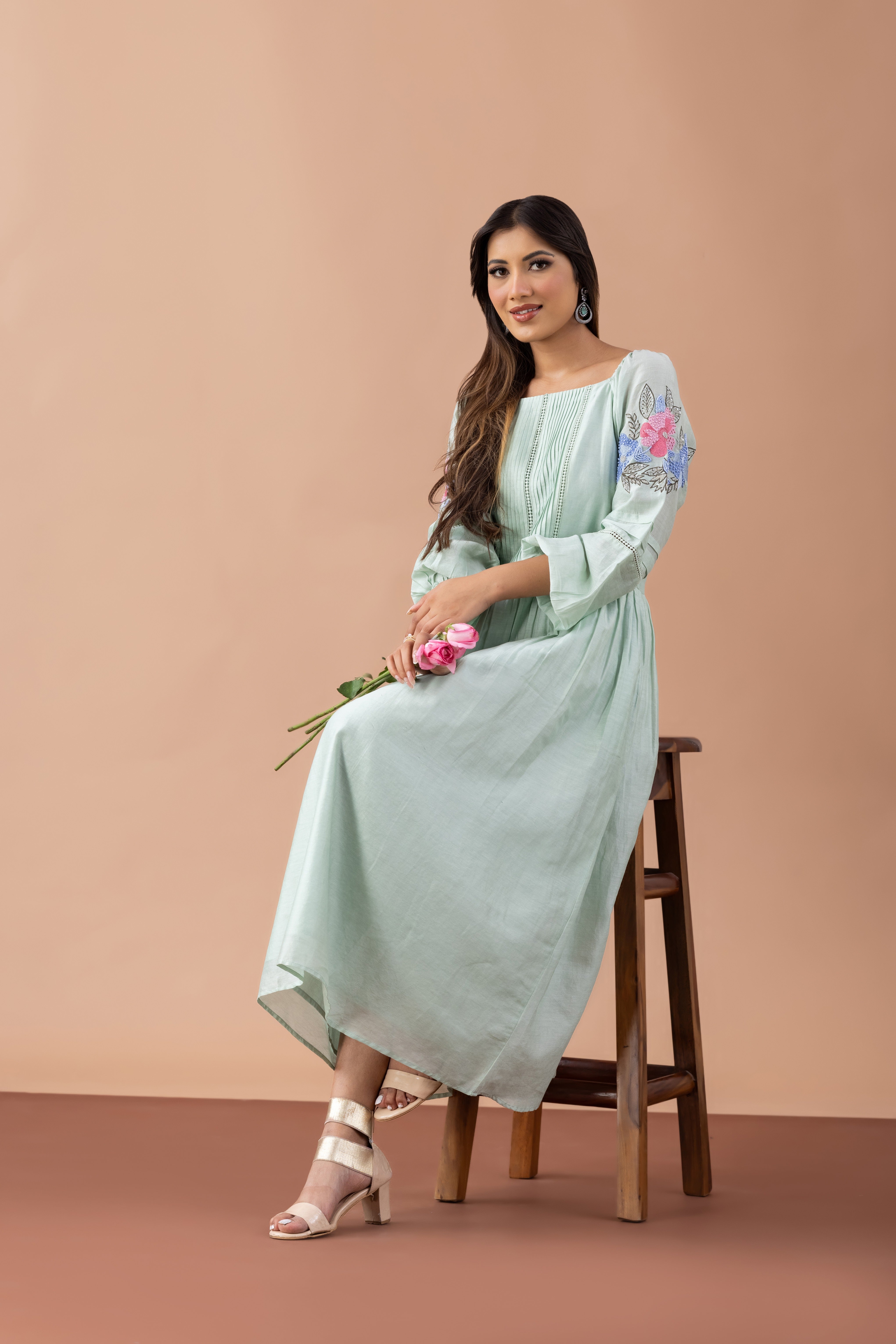 Woman in a light green dress with floral embroidery sitting on a wooden stool against a beige background